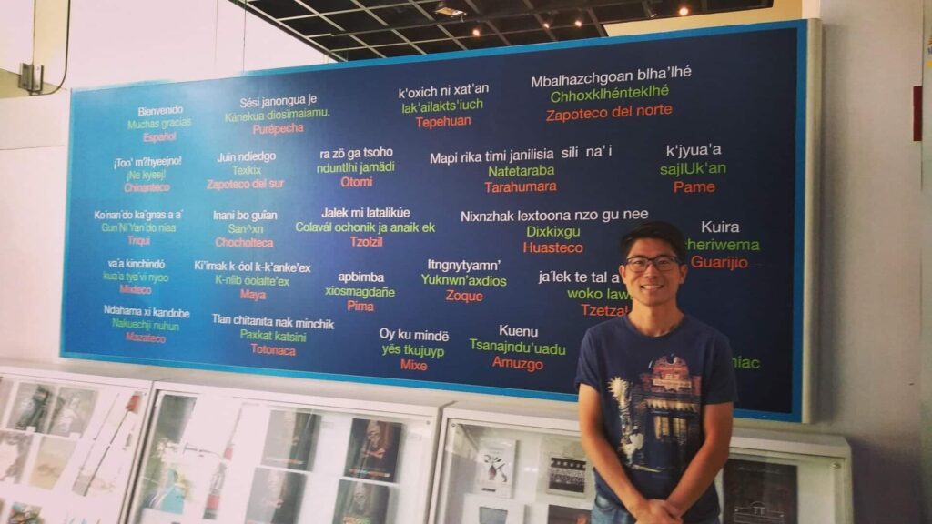 Ko Idioma - メキシコのプライベートツアー | MCT メキシコの観光ガイド岩﨑功がいろいろな言語が書かれたボードの前で挨拶をしている