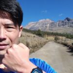 青いシャツを着た男性が険しい山を背景にガッツポーズをしている